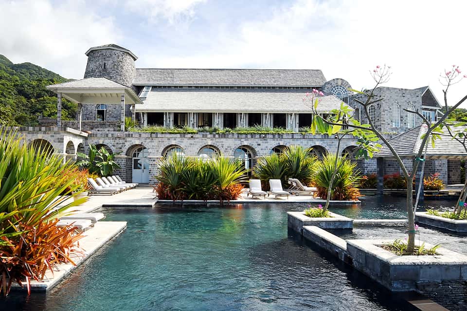Waterfront stone resort building with arched colonnade, lounge chairs, tropical plants, and turquoise water feature