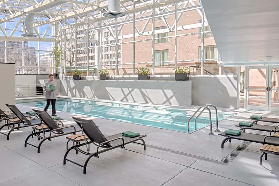 Indoor rooftop pool with floor-to-ceiling windows overlooking urban cityscape and lounge chairs