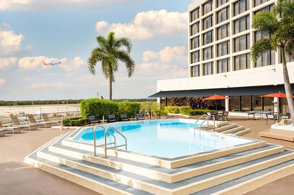 Waterfront resort pool with blue water, lounge chairs, orange umbrellas, palm trees, and bay view