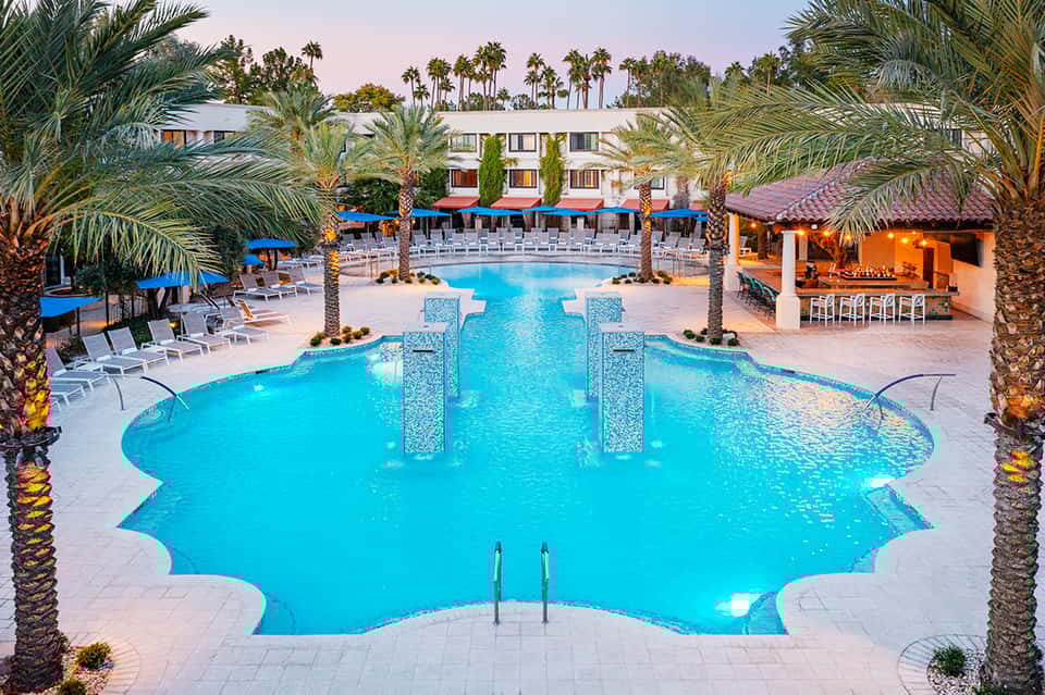 Luxury resort pool with turquoise water, palm trees, and multi-story hotel building with blue umbrellas