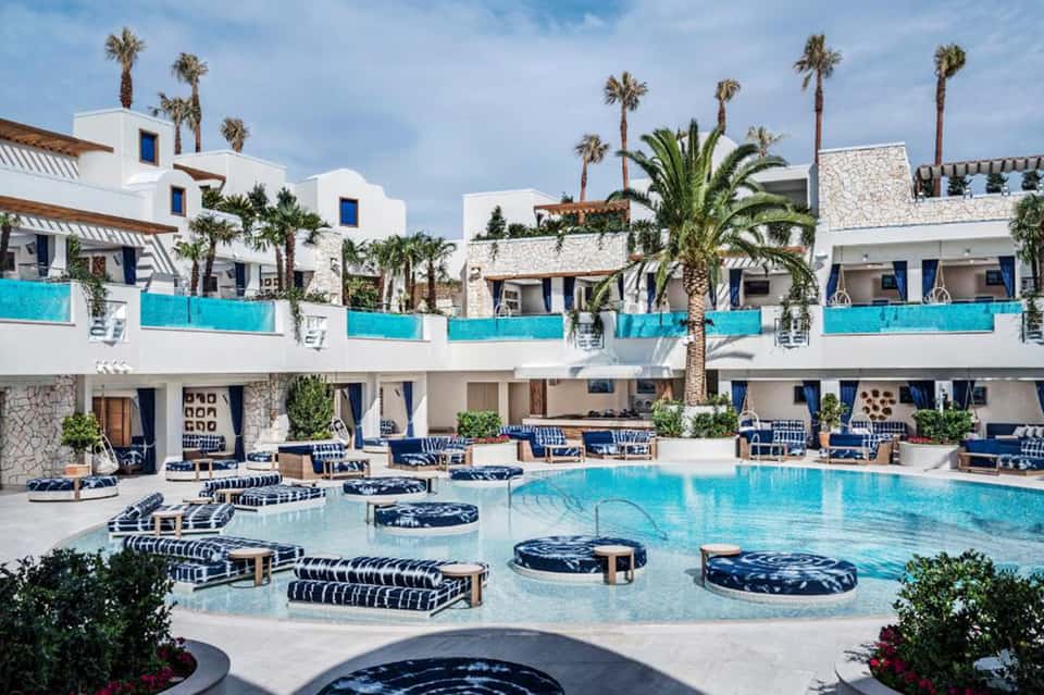Luxury resort courtyard with turquoise pool, white Mediterranean-style buildings, palm trees, and blue loungers