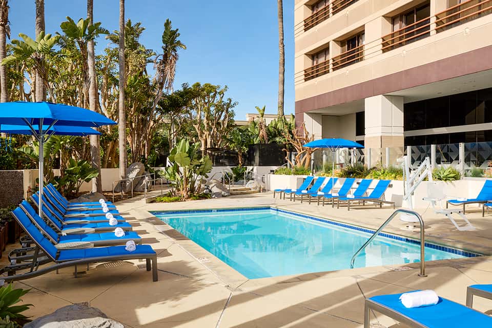 Hotel pool courtyard with blue loungers, palm trees, umbrellas, and multi-story building with balconies under clear sky