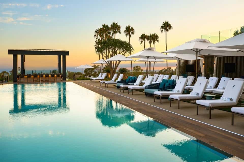 Luxury resort infinity pool at sunset with palm trees, lounge chairs, and cabanas overlooking coastal landscape