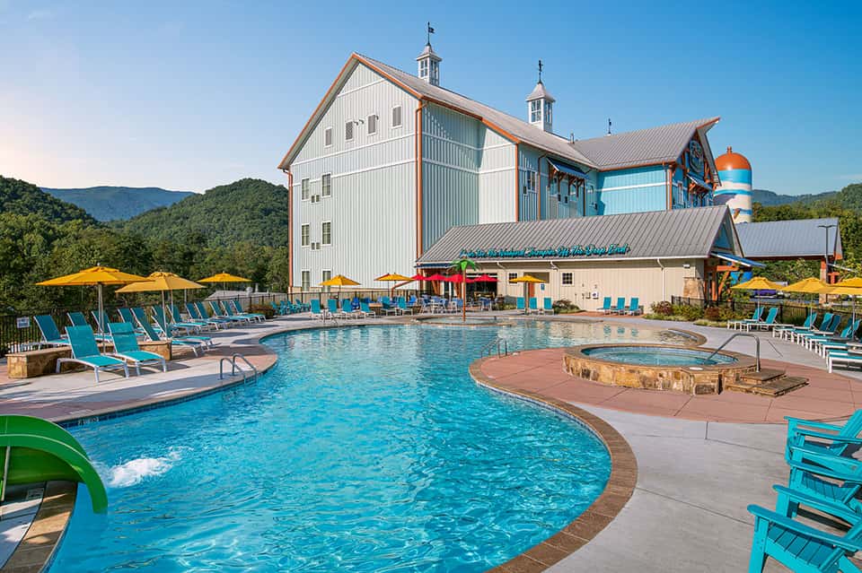 Resort pool area with turquoise water, yellow umbrellas, lounge chairs, and a large barn-style building with mountains in the background