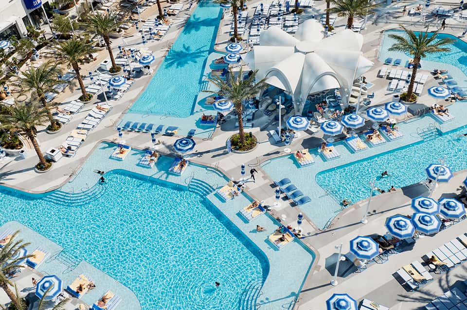Aerial view of resort pools with blue umbrellas, white shade structures, palm trees, and loungers on the deck