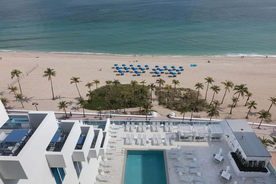 Aerial view of beachfront resort with pool, blue umbrellas, palm trees, and turquoise ocean waters