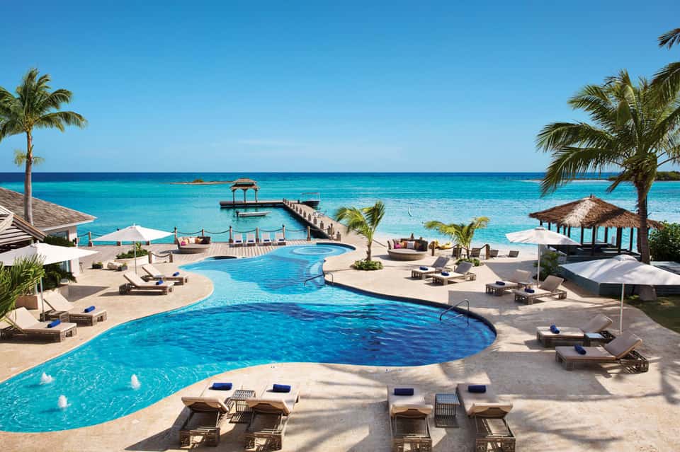 Luxury resort pool area with curved pools, palm trees, ocean view, lounge chairs, and thatched-roof cabanas