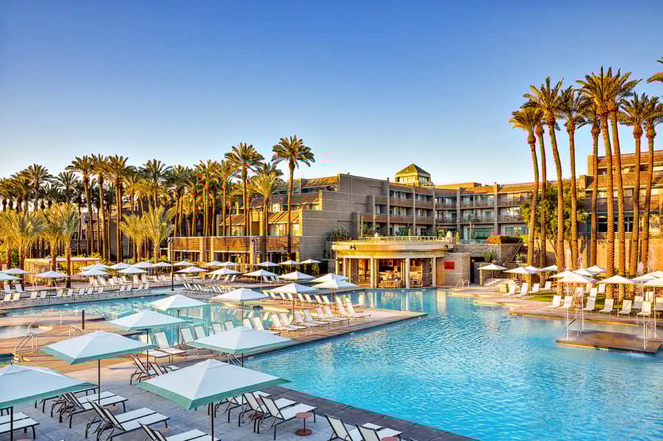 Resort pool area with palm trees, loungers with umbrellas, and multi-story hotel building at sunset