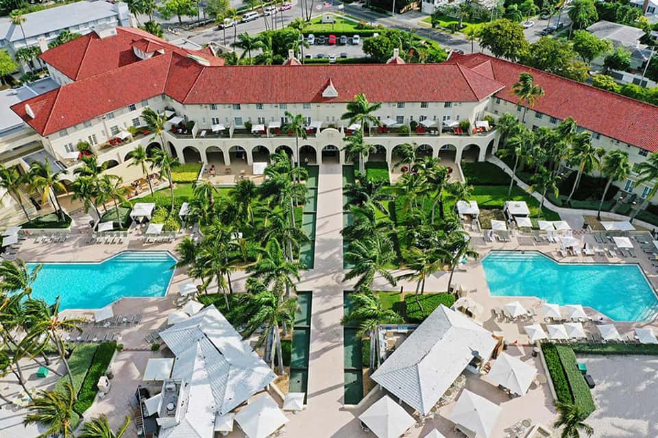 Aerial view of luxury resort with red-roofed buildings, twin pools, palm trees, and manicured grounds