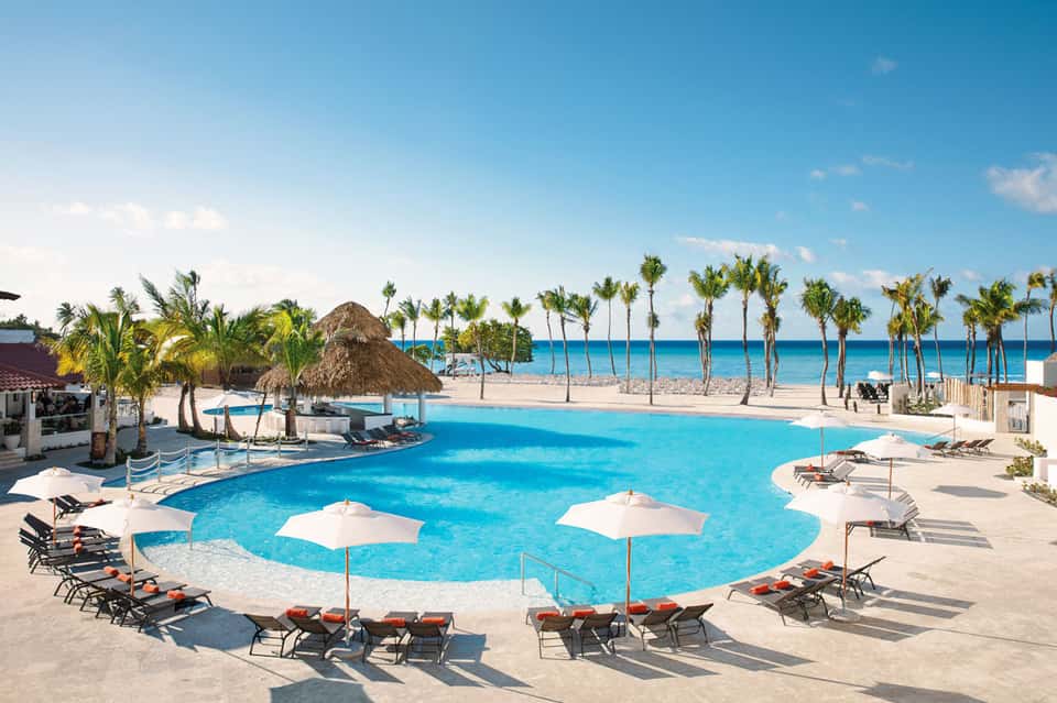Beachfront resort pool with white umbrellas, palm trees, and ocean view with sandy beach in background