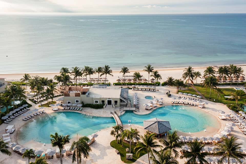 Aerial view of beachfront resort with turquoise pool, palm trees, loungers, and ocean beyond