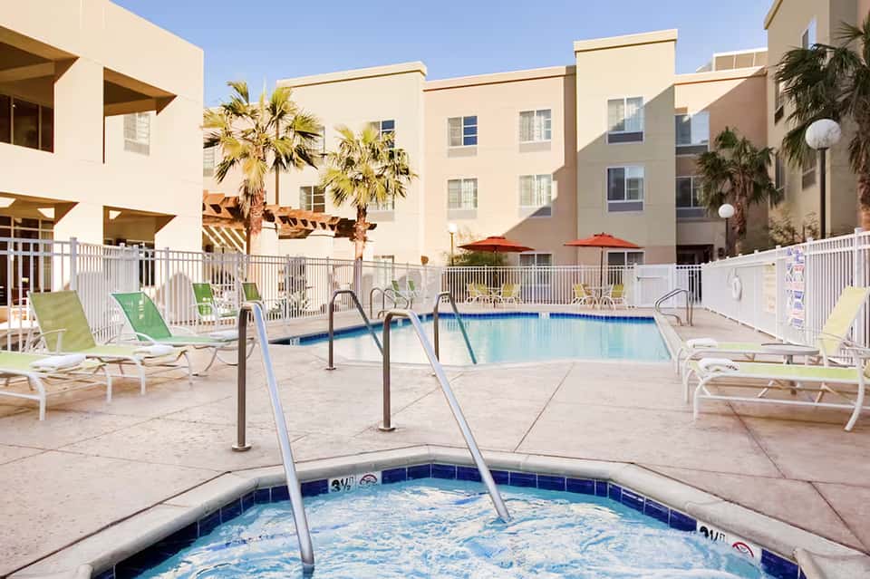 Resort courtyard with swimming pool, lounge chairs, palm trees, and beige apartment buildings under clear sky