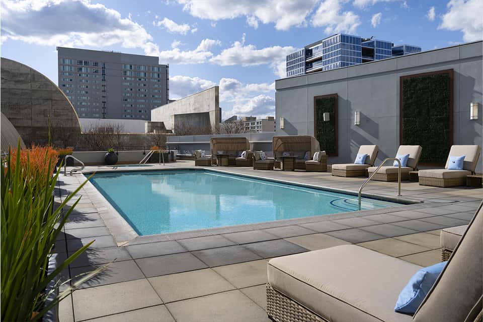 Modern rooftop pool with tan loungers, concrete decking, and city buildings under clear sky