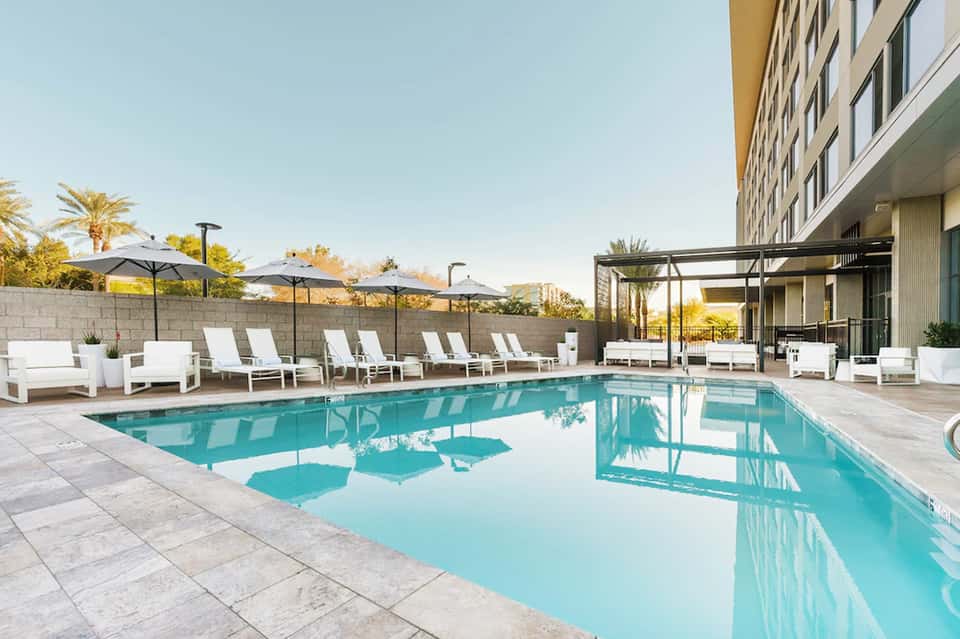 Resort pool deck with turquoise water, white lounge chairs, umbrellas, and high-rise building