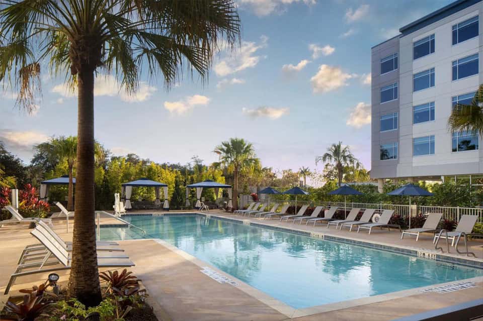 Resort pool with palm trees, lounge chairs, and gazebos beside a modern white hotel building