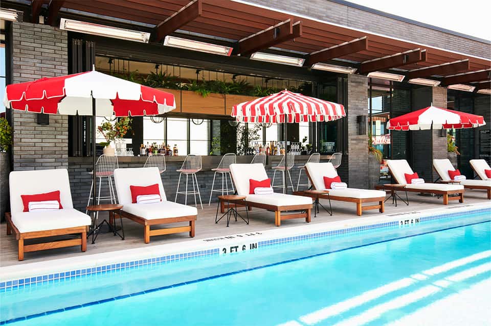 Luxury pool deck with red and white umbrellas, wooden lounge chairs, and modern building with covered patio area