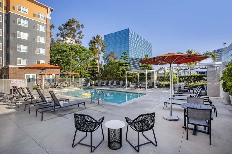 Modern hotel pool deck with loungers, orange umbrellas, contemporary seating areas, and residential buildings