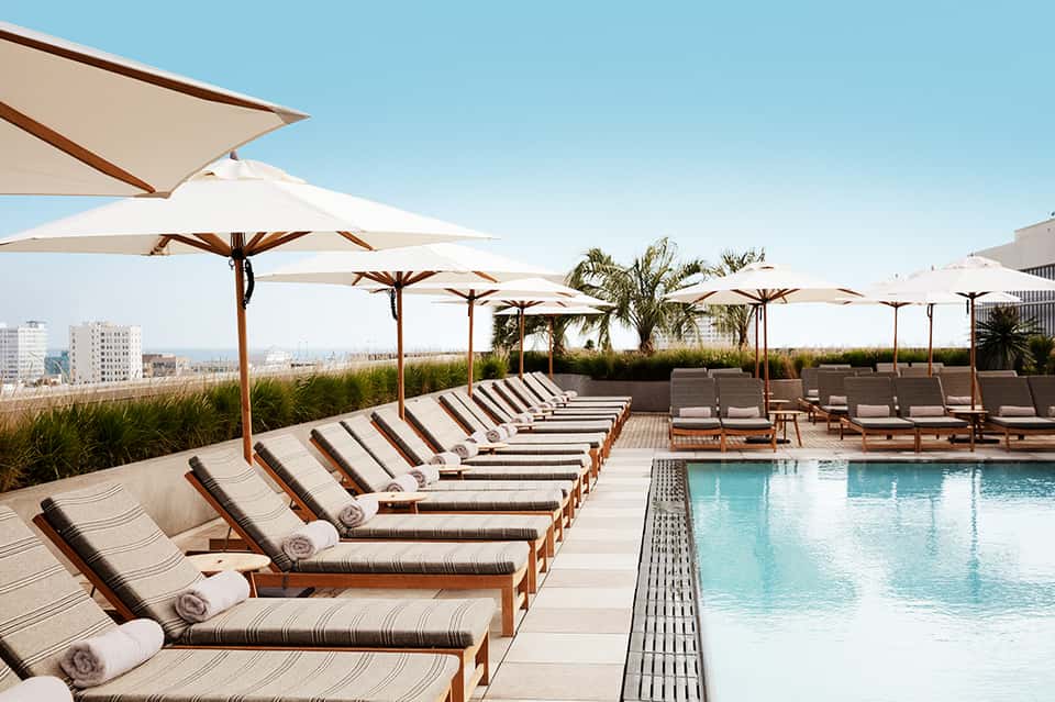 Resort rooftop pool deck with lounge chairs, umbrellas, palm trees, and city skyline at sunset