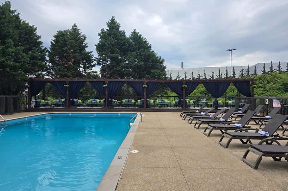 Resort pool with crystal blue water, black lounge chairs, and covered pergola with dark drapes overlooking mountains