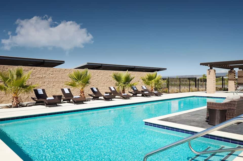 Modern resort pool with turquoise water, black lounge chairs, stone wall, and palm trees under blue sky