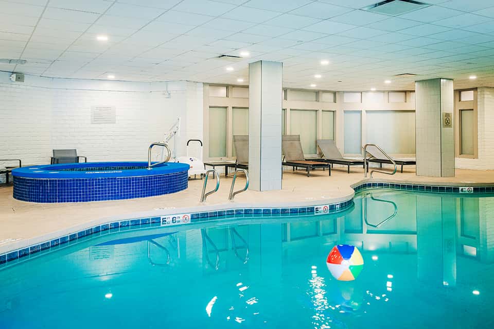 Indoor pool facility with blue water, hot tub, lounge chairs, and columns