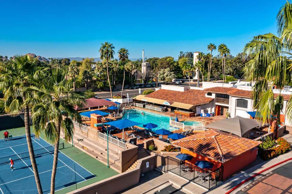 Resort pool with blue water, palm trees, tennis courts, and desert mountains in the background under clear sky