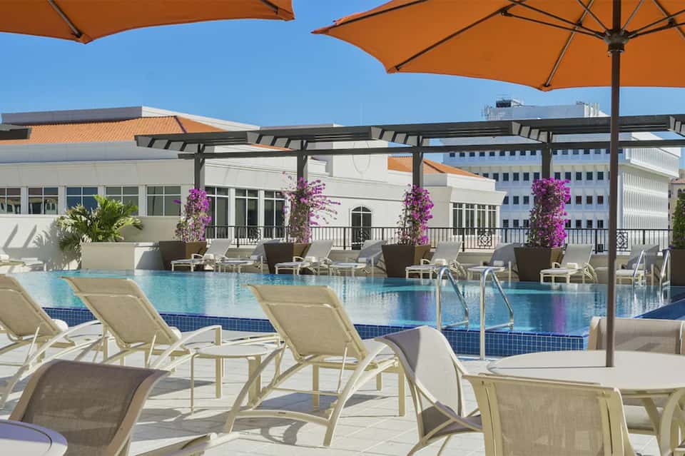 Resort pool deck with orange umbrellas, lounge chairs, pergola with purple hanging flowers, and modern buildings