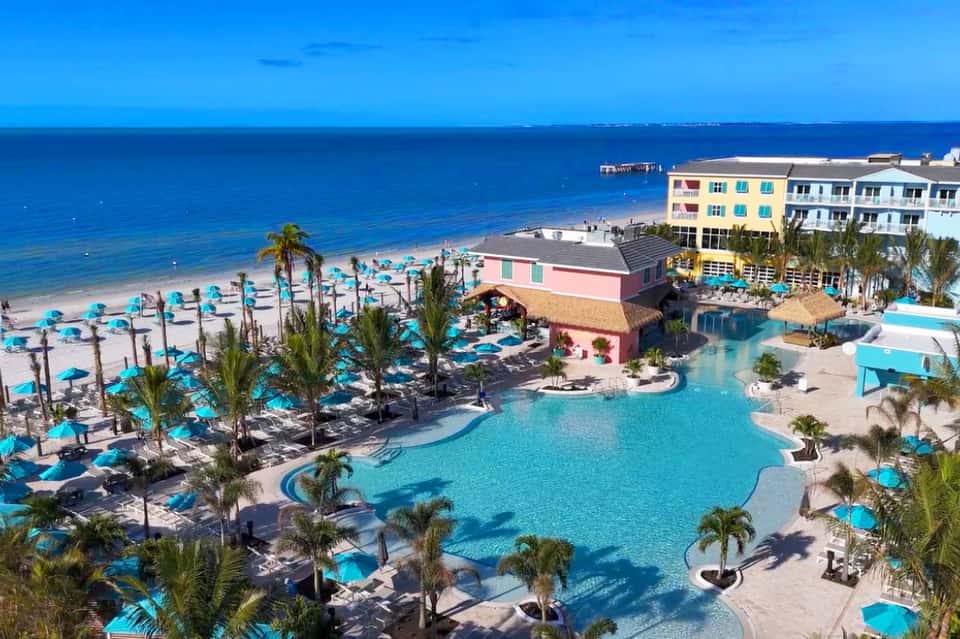 Aerial view of beachfront resort with turquoise pool, palm trees, and colorful umbrellas along sandy beach