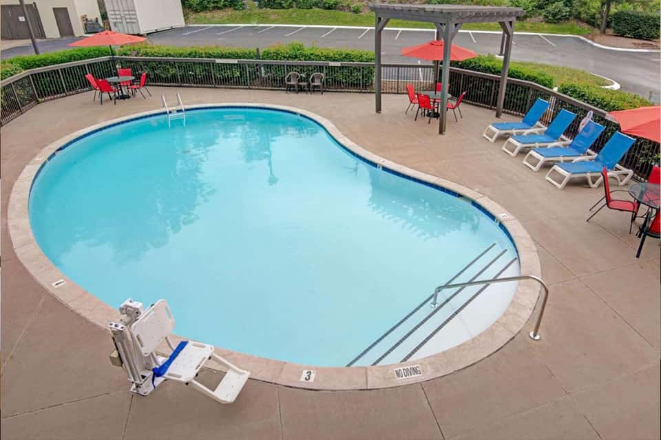 Curved swimming pool surrounded by concrete deck with red umbrellas, pergolas, and blue lounge chairs