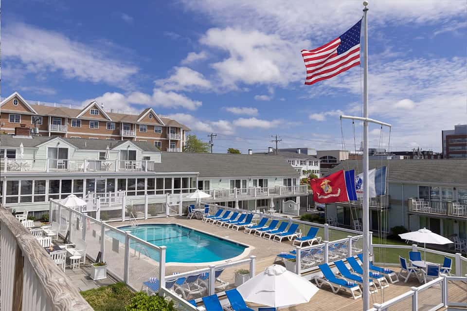 Beachfront resort pool with blue loungers, American flag, and coastal buildings under clear sky