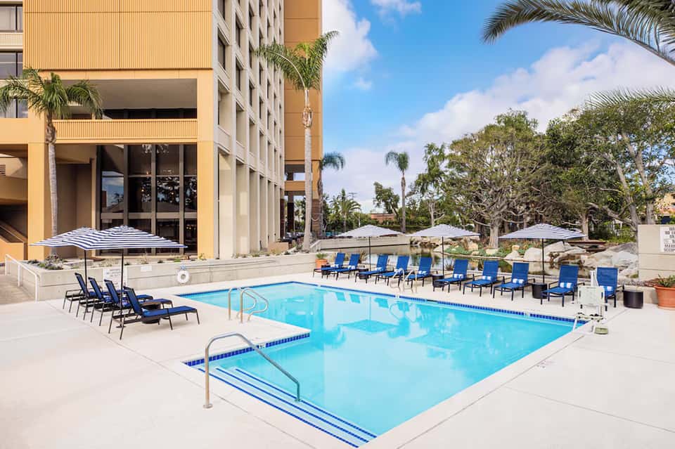Resort outdoor pool with blue water, loungers, umbrellas, and multi-story building with palm trees