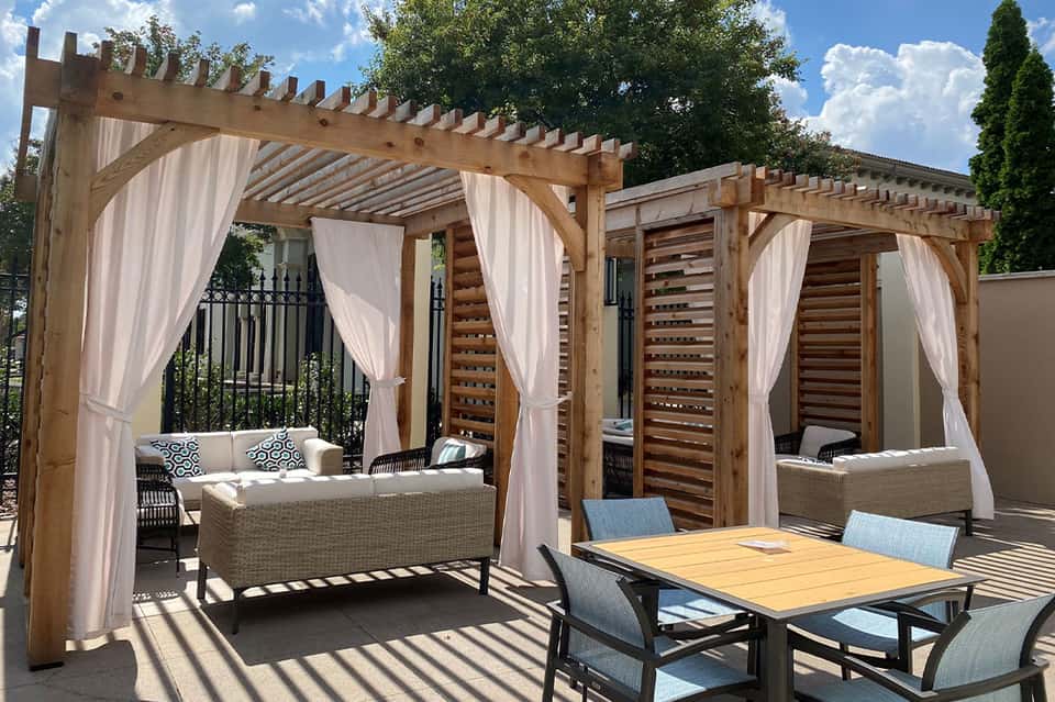 Wooden pergola with white curtains, wicker seating, dining table, and shade structures on outdoor patio