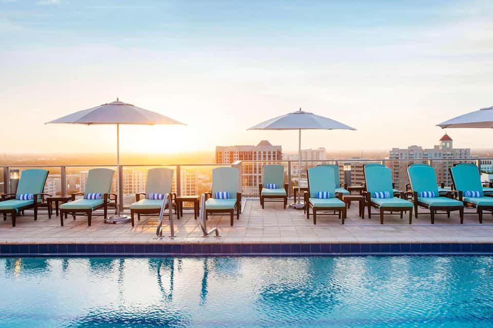 Rooftop pool deck at sunset with turquoise loungers, umbrellas, and city skyline views