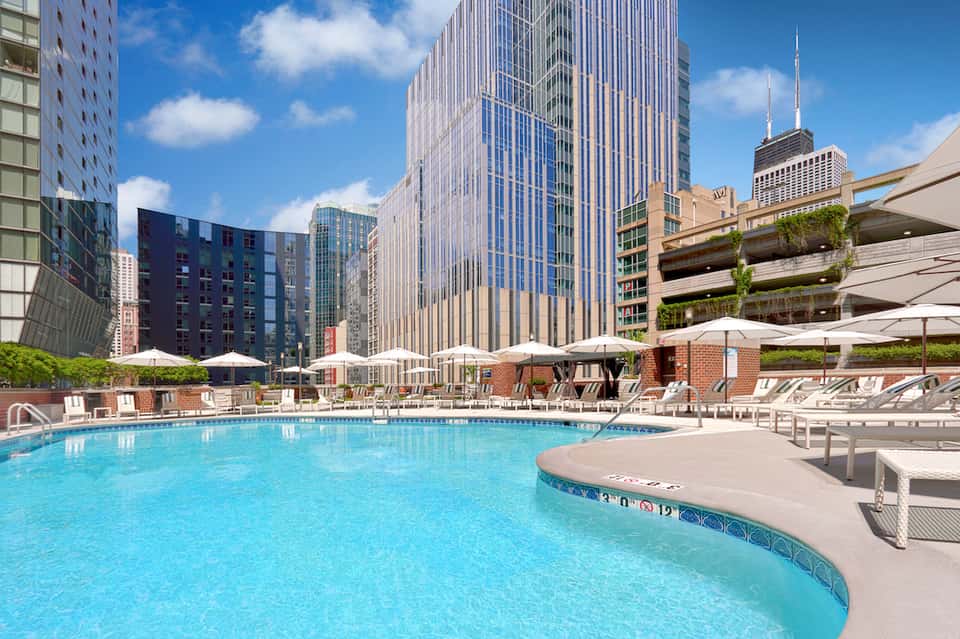 Urban rooftop pool surrounded by downtown skyscrapers, white umbrellas, and lounge seating