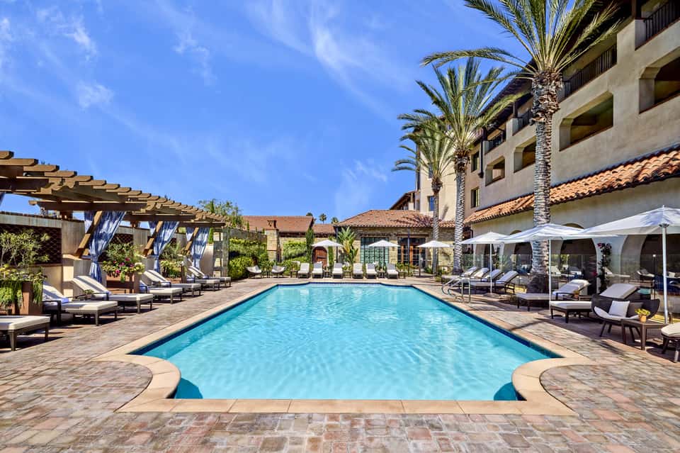 Rectangular lap pool surrounded by lounge chairs, pergolas, and Spanish colonial architecture