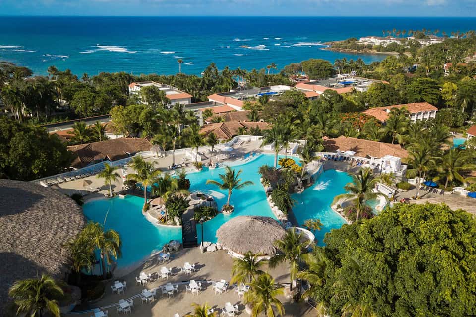 Aerial view of resort with large lagoon-style pool, palm trees, and ocean coastline