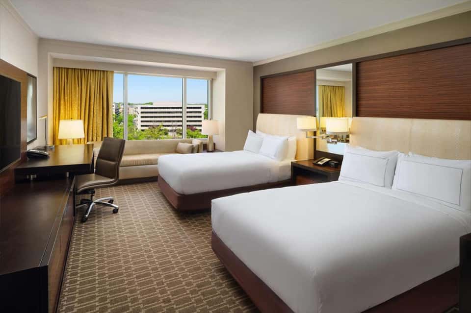 Hotel room with two queen beds, dark wood headboards, golden curtains, modern decor, and city view windows