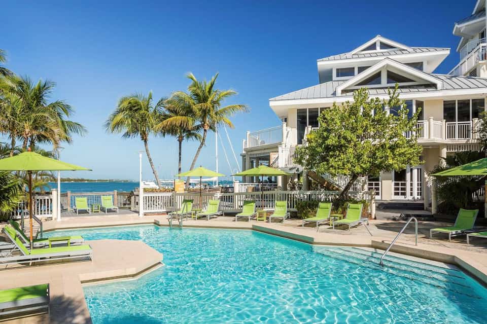 Beachfront resort with pool, palm trees, white umbrellas, lounge chairs, and sailboat in background