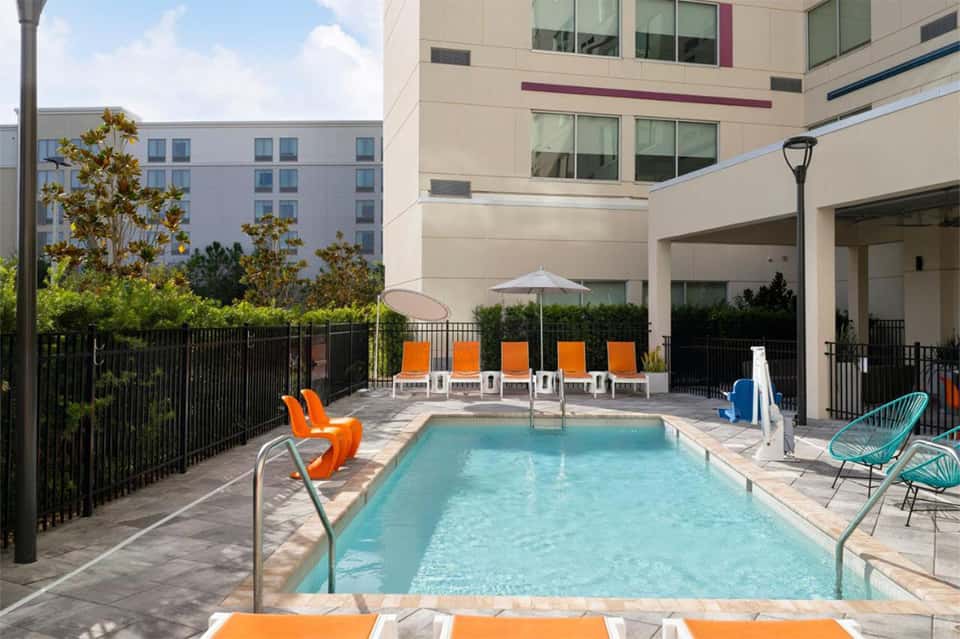 Outdoor pool deck with turquoise water, orange lounge chairs, black fencing, and multi-story hotel building