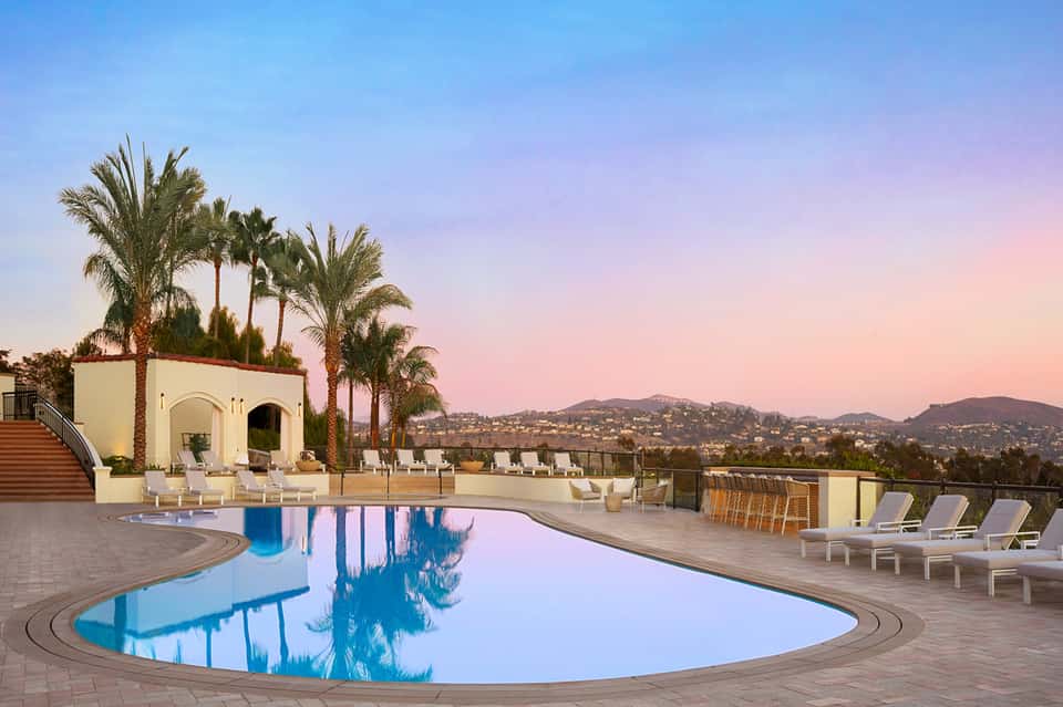 Luxury resort pool at dusk with palm trees, lounge chairs, and hillside views at sunset