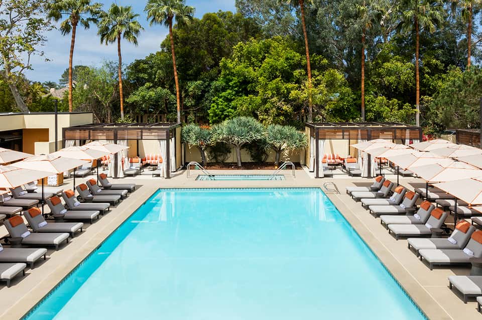Resort pool surrounded by lounge chairs, white umbrellas, palm trees, and lush landscaping during daytime
