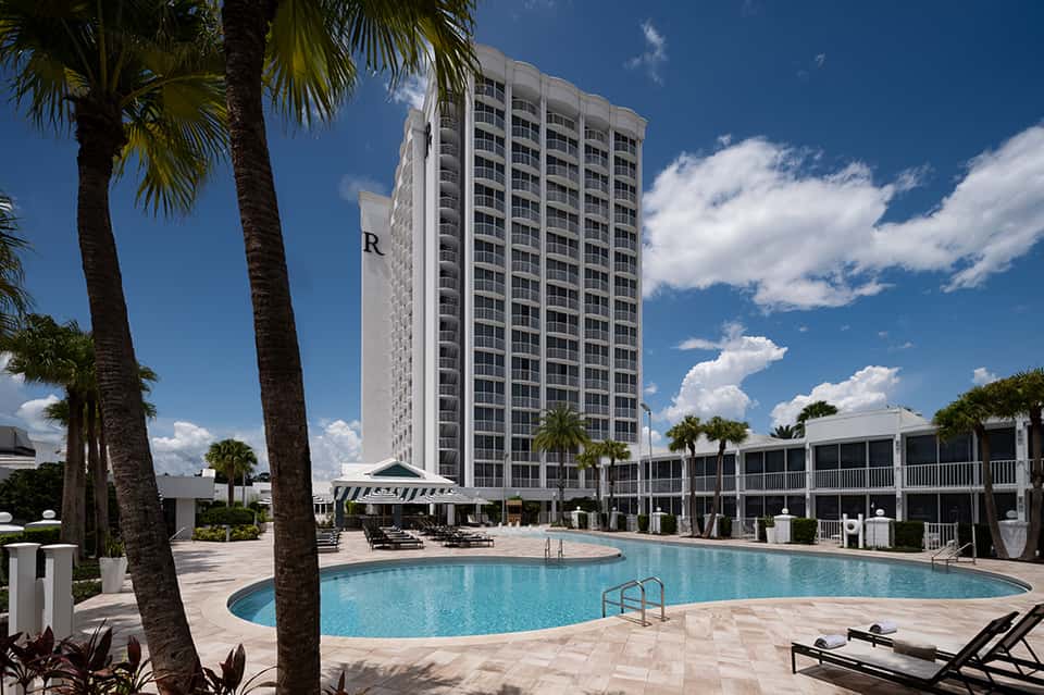 Beachfront resort exterior with palm trees, multi-story hotel tower, and curved swimming pool under blue sky