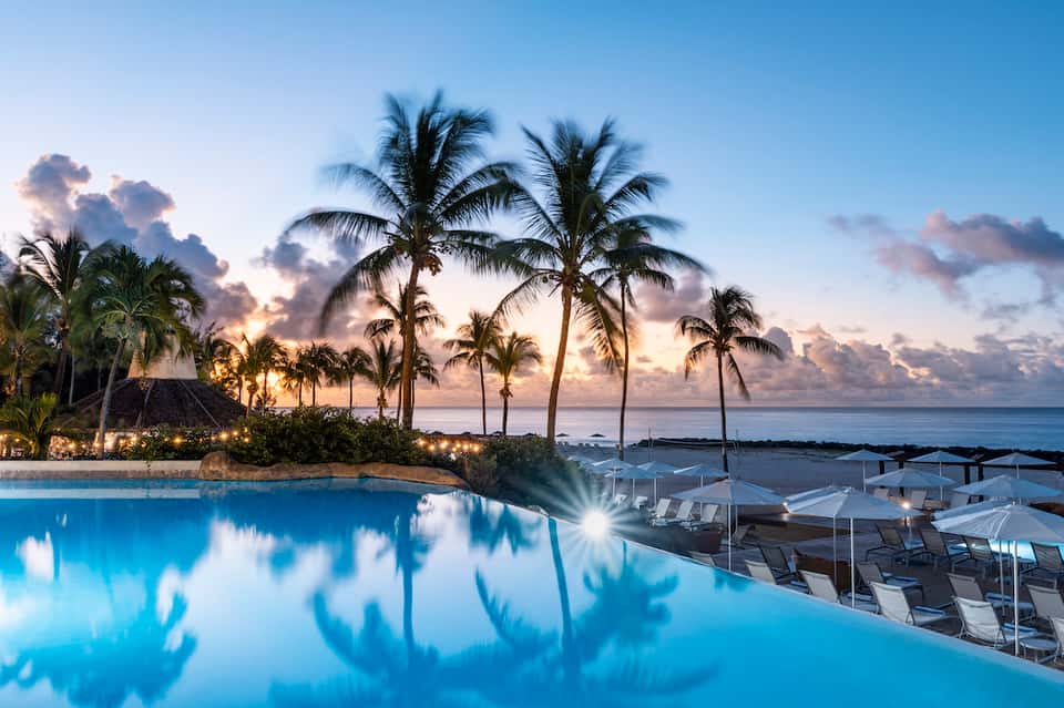 Illuminated infinity pool at dusk with palm trees, lounge chairs, and ocean horizon