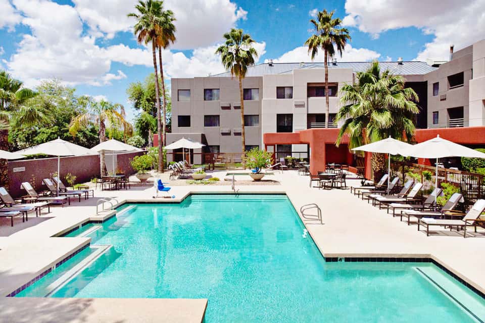Modern resort courtyard with rectangular pool, palm trees, and contemporary multi-story hotel buildings with white umbrellas