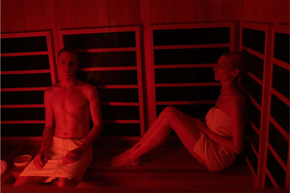 Two people relaxing in red-lit sauna with wooden benches and horizontal slats