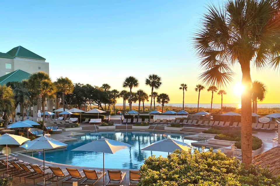 Resort pool at sunset with palm trees, lounge chairs, umbrellas, and beachfront buildings with ocean views