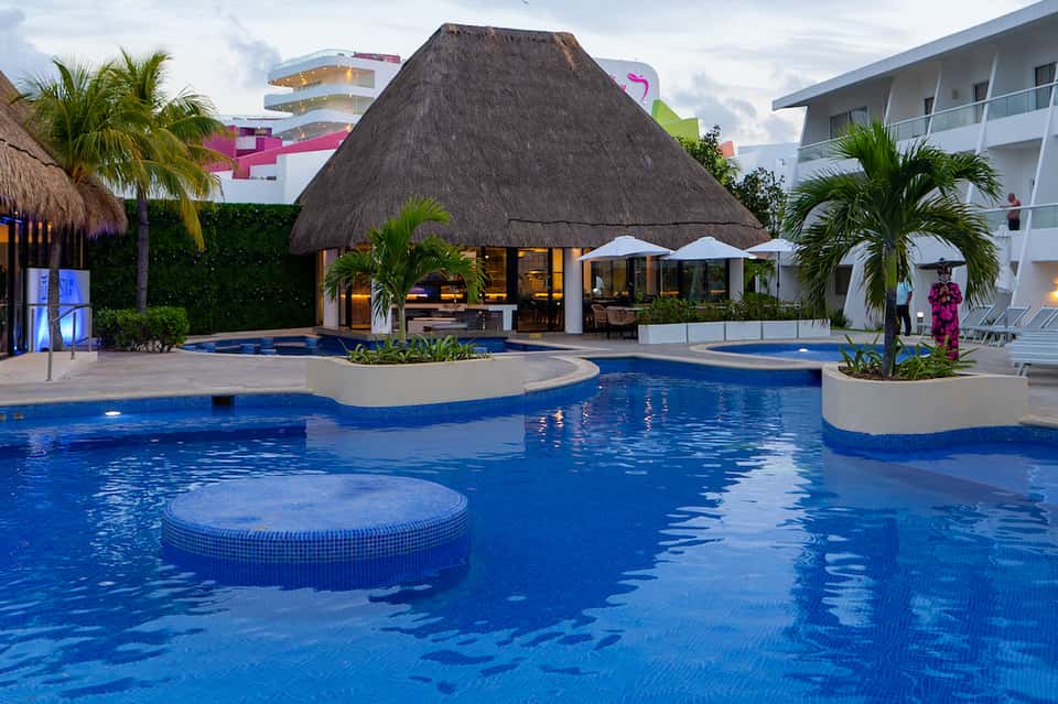 Resort pool complex with thatched-roof bar, palm trees, and modern hotel buildings