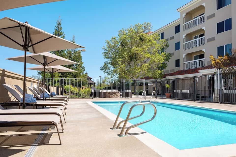 Resort swimming pool with white loungers, beige umbrellas, and multi-story building backdrop