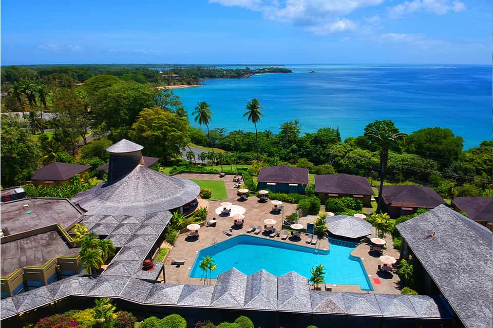 Aerial view of tropical resort with pool, thatched-roof bungalows, palm trees, and turquoise ocean coastline
