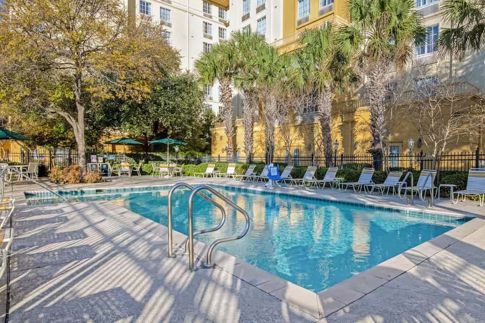 Resort pool with green umbrellas, lounge chairs, and palm trees surrounded by yellow buildings